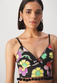 Čierny crop top s farebnými kvetinovými vzormi, nastaviteľnými ramienkami a trimom s pom-pom na spodnom leme. Textúrovaná látka.
