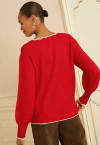 Roter Strickpullover mit gewelltem Ausschnitt und weißen Akzenten, ausgestattet mit langen, puffigen Ärmeln und einem strukturierten Stoff. Kombiniert mit braunen Hosen.