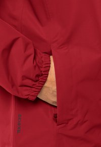 Giacca da esterno rossa con polsini elasticizzati e tasca laterale. Il materiale sembra liscio con una finitura opaca. Marchio "TEXIPORE" visibile.
