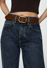 Jeans de mezclilla azul oscuro con un cinturón de cuero marrón y hebilla de latón. Los jeans tienen un corte recto y un diseño de cinco bolsillos.