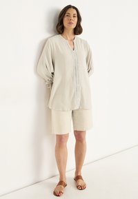 Vrouw staat tegen de muur in een beige tuniek met kantdetails, bijpassende shorts en bruine sandalen met roze teennagels.