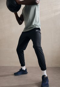 Athlet in einem hellgrünen, kurzärmeligen Shirt, schwarzen, taillierten Hosen und blauen New Balance Schuhen, der einen schwarzen Medizinball hält.
