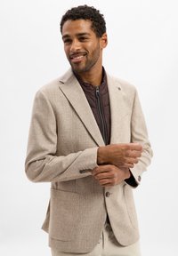 Mann lächelt, passt ein cremefarbenes karierter Blazer über eine braune gesteppte Zip-Jacke an, steht vor einem einfachen weißen Hintergrund.