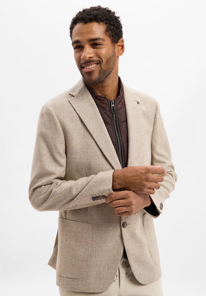 Mann lächelt, passt ein cremefarbenes karierter Blazer über eine braune gesteppte Zip-Jacke an, steht vor einem einfachen weißen Hintergrund.