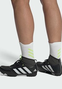 Baskets de sport noires avec des rayures blanches, dotées d'une chaussette de cheville en maille texturée grise avec des accents vert fluo. Conçues pour la performance.