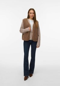 Brown vest with plush texture, beige sleeves, and button closures, worn over a white top and paired with dark flared jeans and brown heels.