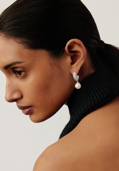 Γυναίκα με σκούρα μαλλιά σε χαμηλή αλογοουρά, που φοράει μαύρο πλεκτό αμάνικο τοπ και ασημένια σκουλαρίκια με σταγόνες από μαργαριτάρι, κοιτάζει προς τα κάτω αριστερά.