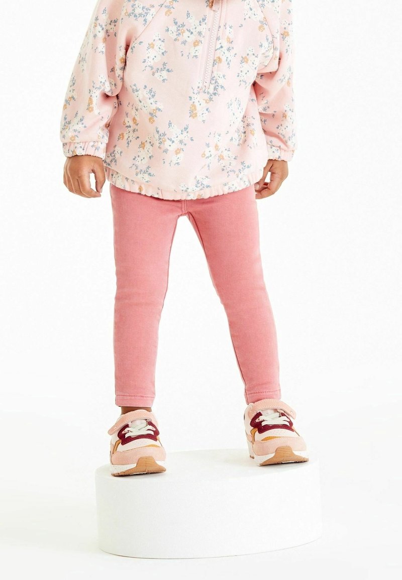 Enfant portant une veste fleurie rose, des leggings roses et des baskets roses, debout sur une plateforme ronde blanche sur fond blanc.