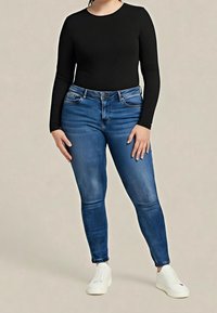 Personne portant un haut noir ajusté à manches longues, un jean skinny bleu et des baskets blanches, debout devant un fond uni.