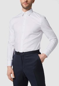 Weißes Langarmhemd mit Button-Down-Kragen, figurbetontem Design, glatter Textur, kombiniert mit marineblauen Hosen mit Gürtelschlaufen.