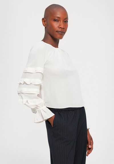 Femme chauve et confiante portant un chemisier blanc avec des manches transparentes à volant et un pantalon à rayures navy, posant devant un fond uni.