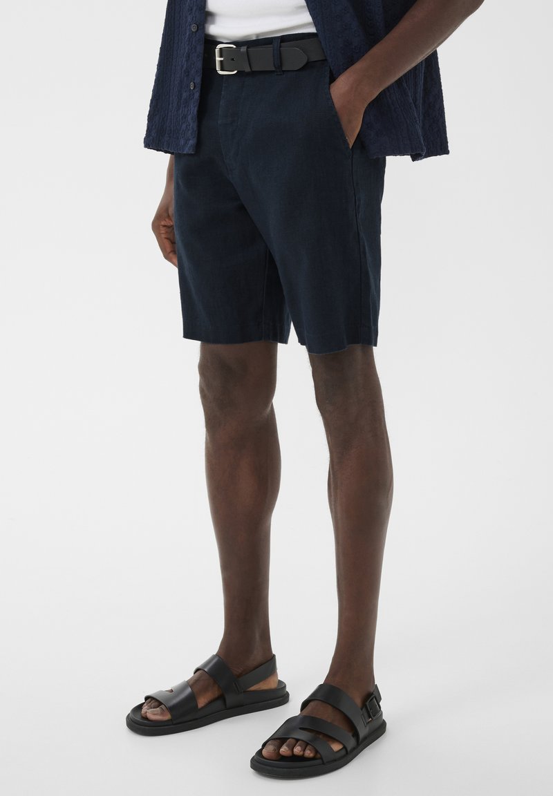 Homme portant un short bleu marine, une ceinture noire, une chemise bleu marine et des sandales noires à bouts ouverts, debout avec une main dans la poche.