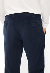 Pantaloni in corduroy blu navy con una superficie testurizzata, due tasche posteriori con bottoni e una vestibilità slim. Camicia bianca visibile in vita.