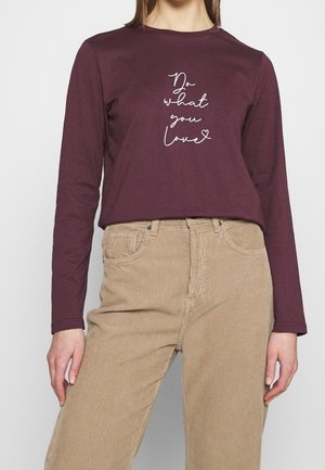 Femme portant une chemise à manches longues bordeaux avec le texte « Fais ce que tu aimes » et un pantalon en velours beige, debout devant un fond uni.
