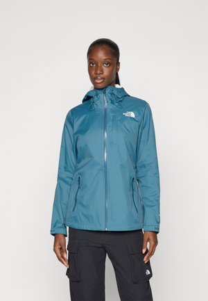 Veste imperméable teinte sarcelle avec capuche, fermeture éclair avant, cordon de serrage réglable et logo sur la poitrine ; associée à un pantalon cargo.