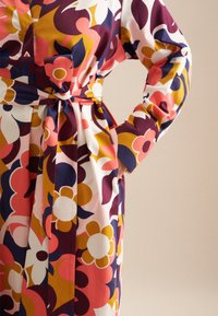 Buntes Blumenkleid mit einem lebhaften Muster in Pink, Gelb und Marineblau, mit einem taillierten Gürtel und langen Ärmeln. Aus weichem Stoff hergestellt.