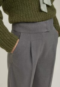 Pantaloni grigi su misura con una texture liscia e frontale plissettato, abbinati a un maglione in maglia verde oliva morbido che mostra una vestibilità rilassata.