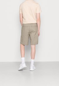 Persoon in een beige korte broek, lichtbeige t-shirt, witte crew-sokken en witte sneakers staat met het gezicht naar een effen witte muur.