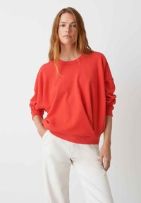 Sweatshirt rouge surdimensionné avec un col rond et des manches longues, associé à un pantalon de couleur claire. Texture lisse et coupe décontractée.