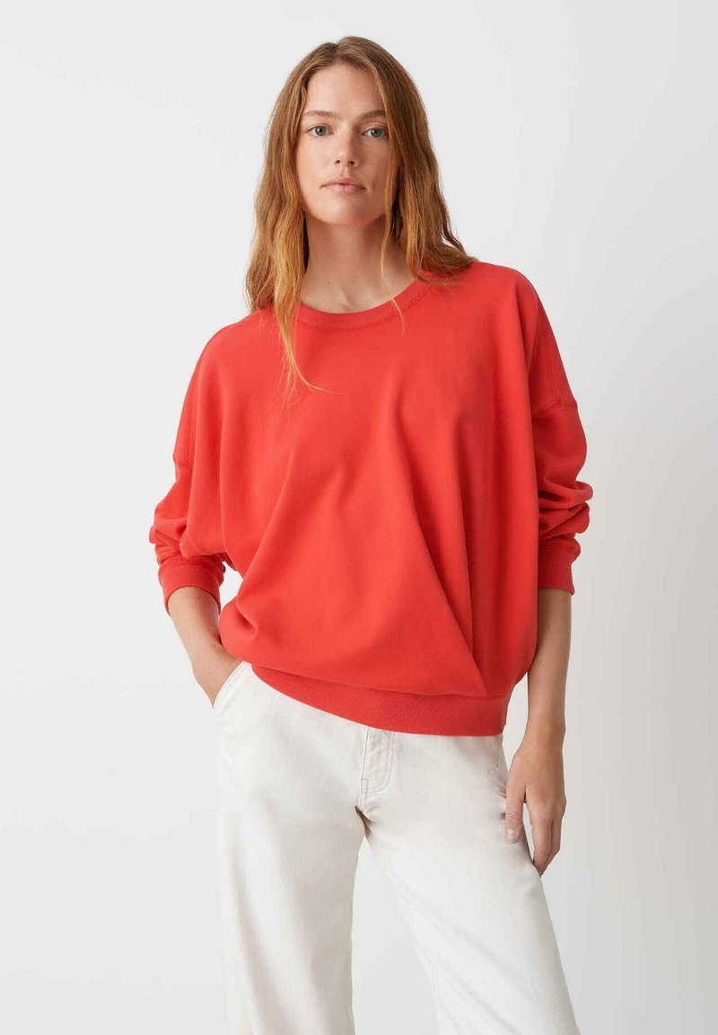 Sweatshirt rouge surdimensionné avec un col rond et des manches longues, associé à un pantalon de couleur claire. Texture lisse et coupe décontractée.
