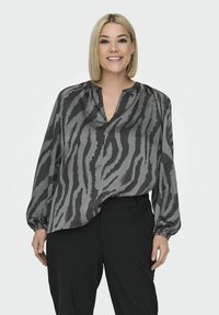 Grå och svart blus med tigerstrimmigt mönster, V-ringning och långa ärmar; matchad med svarta byxor. Mjuk, sidenliknande textur.