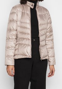 Pružná bunda z lesklého béžového saténového materiálu, vybavená vysokým límcem, zipovým zapínáním a vodorovným prošívaným vzorem. Měkká textura.