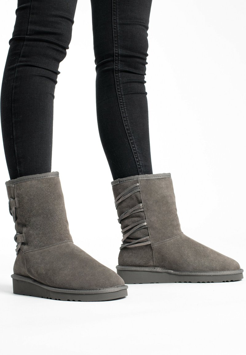 Stivaletti alla caviglia in suede grigio con punta arrotondata, suola piatta e lace decorativi sui lati. Indossati con jeans skinny neri, caratterizzati da una texture liscia.