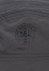 Grauer Stoff mit einem subtilen Karomuster, das ein kreisförmiges, in Schwarz eingeprägtes Logo zeigt. Strukturelle Nähte sind sichtbar.