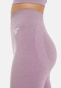 Lila leggins visokim pasom iz elastičnega materiala, z bočno žepom in belim logotipom na boku. Gladka tekstura.