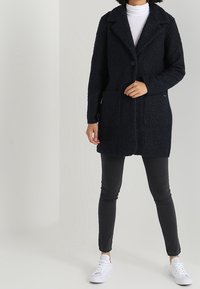 Mujer vestida con un abrigo negro texturizado, un suéter blanco de cuello alto, jeans skinny negros y zapatillas blancas, de pie contra un fondo claro y liso.