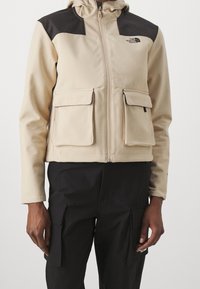 Chaqueta beige con cremallera y acentos negros en los hombros, dos bolsillos delanteros con solapa, tela suave, capucha ajustable y logotipo en el lado izquierdo del pecho.