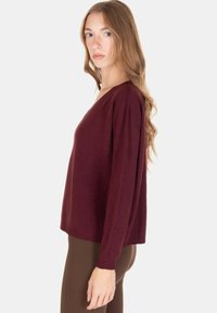 Giovane donna con lunghi capelli mossi, che indossa un maglione bordeaux largo e pantaloni marroni, in piedi di profilo contro uno sfondo bianco.