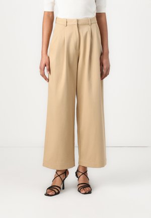 Beige vidde bukser med frontfolder, høj talje og en glat tekstur. Sammen med sorte strophæle.