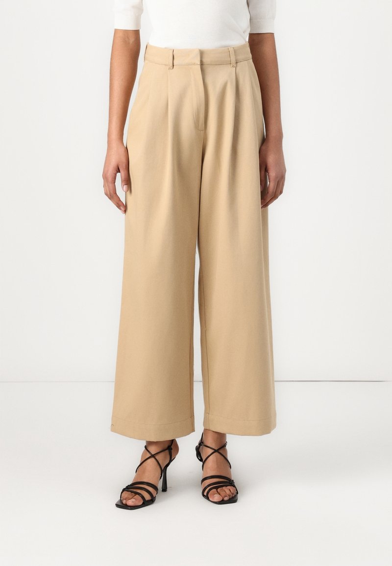 Beige vida byxor med frontveck, hög midja och en slät textur. I kombination med svarta remsandaler.