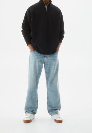 Person trägt schwarzen Viertel-Reißverschluss-Sweatshirt, hellblaue Jeans mit weitem Bein, weiße Sneaker mit beige farbenen Sohlen, steht mit den Händen in den Taschen.