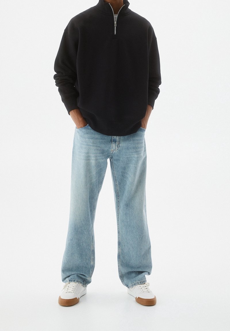 Personne portant un sweat-shirt noir à fermeture quart de zip, un jean bleu clair à jambes larges, des baskets blanches avec des semelles beige, debout les mains dans les poches.