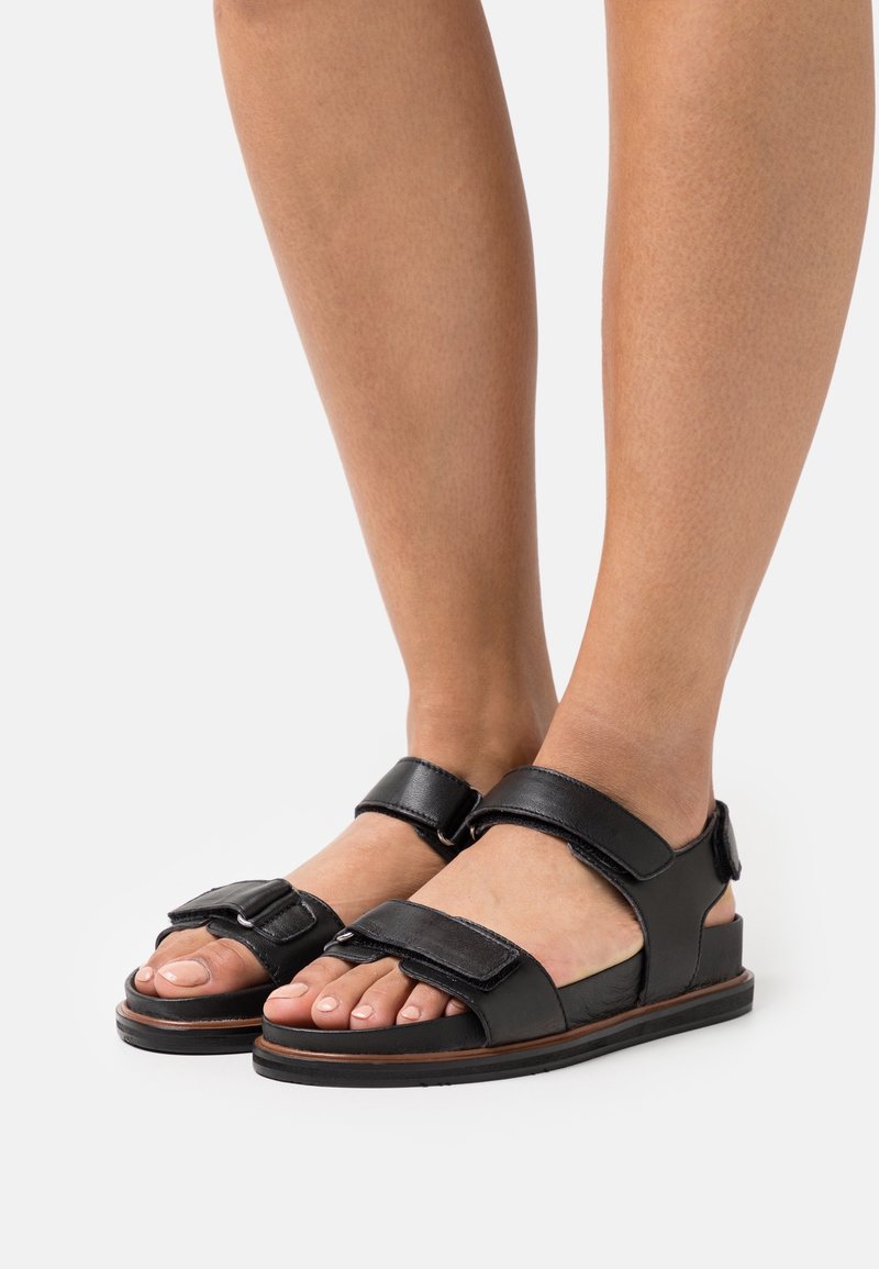 Sandales noires à bout ouvert avec deux brides réglables, une semelle plate et une finition en cuir lisse. Accent métallique notable sur la bride avant.