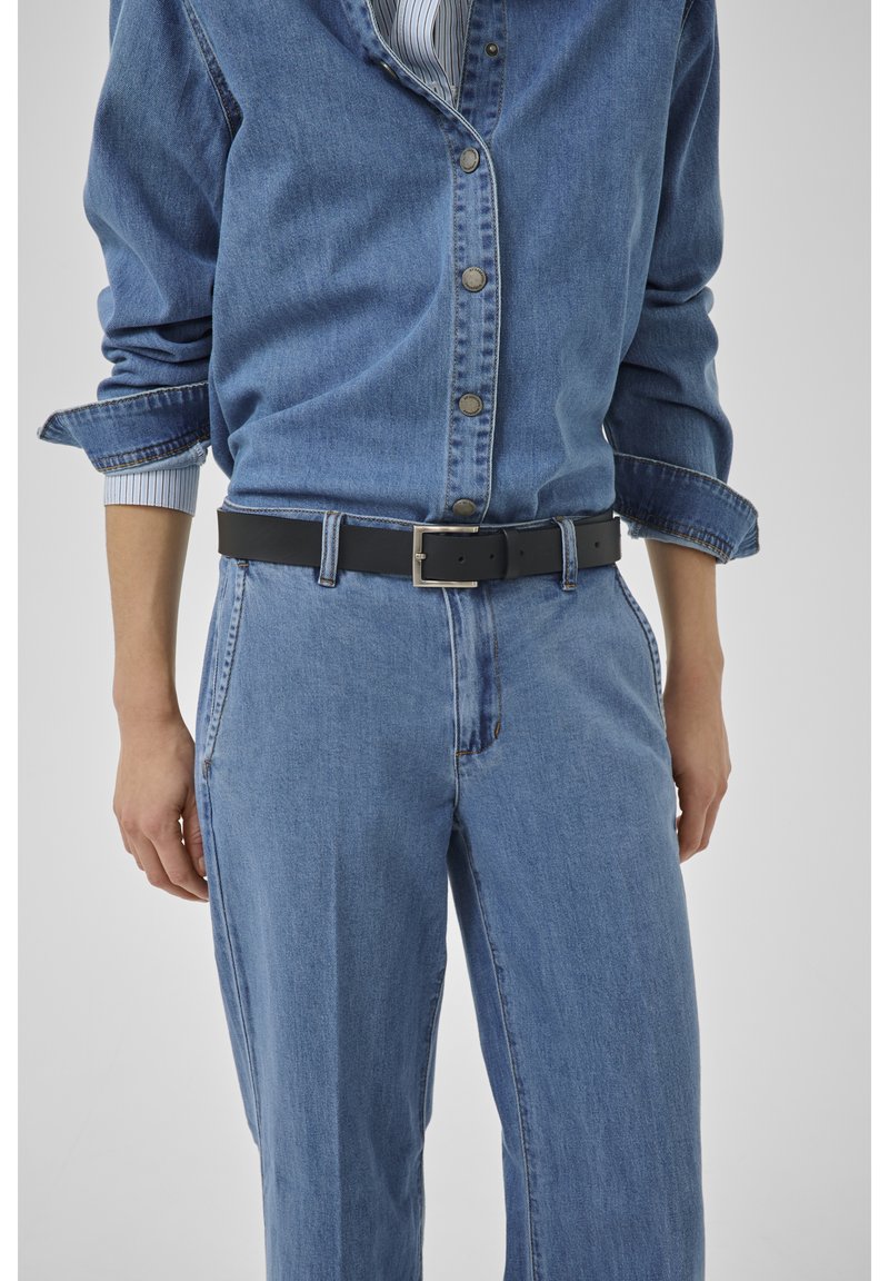 Henkilö, jolla on sininen farkkupaita ja farkut, hihat käännettyinä ylös, sekä musta vyö hopeisella soljella, seisomassa yksiväristä taustaa vasten.