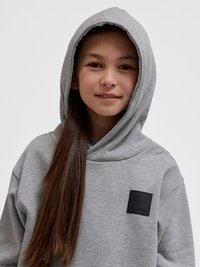 Fille aux longs cheveux bruns portant un sweat à capuche gris avec un patch logo noir Hummel, souriante sur un fond clair uni.
