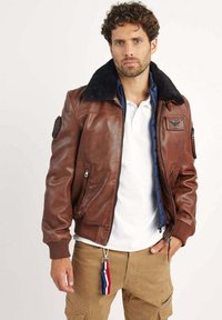 Veste en cuir marron de style bombardier avec un col en fourrure noire, fermeture éclair à l'avant et poches latérales. Présente un patch sur le bras gauche et des poignets côtelés.