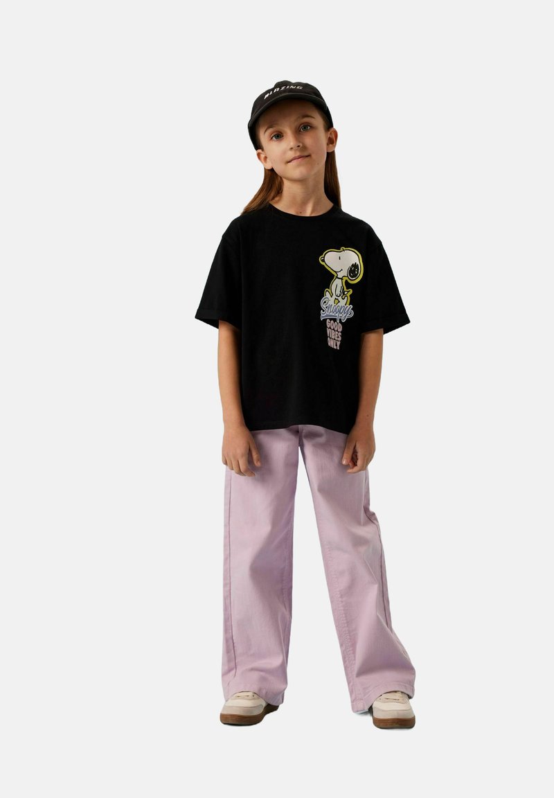 Musta lyhythihainen T-paita, jossa on Snoopy-kuvio, yhdistetty vaaleanpinkin leveälahkeisten housujen ja valkoisten tennareiden kanssa, neutraalilla taustalla.