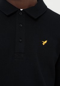 Μαύρο πόλο με γιακά, τρία κουμπιά και μικρό κίτρινο κεντημένο λογότυπο πουλιού στο αριστερό μέρος του στήθους.