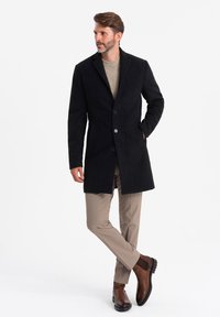 Abrigo de lana negra con un corte entallado, diseño de un solo botón, dos bolsillos frontales y botones redondos, combinado con pantalones beiges y botas marrones.