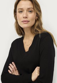 Black v-neck sweater with a fine knit texture, featuring long sleeves. Minimal gold jewelry accents. Background is plain and light.