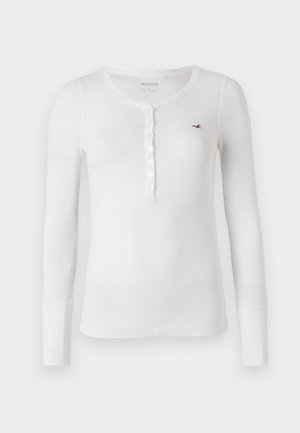 Cămiză albă cu mânecă lungă tip Henley, cu textură ribbed și placket cu nasturi. Prezintă un mic logo roșu pe pieptul stâng și un guler rotunjit.