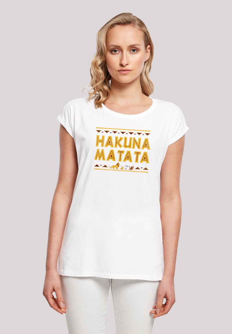 Valkoinen puuvillapaita, jossa on "HAKUNA MATATA" keltaisilla kirjaimilla, ympäröitynä oransseilla ja ruskeilla heimokuvioilla. Lyhyet hihat ja pyöreä kaulus.