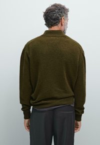 Oude man met grijs haar en baard, gekleed in een olijfgroene gebreide trui en donkere broek, met zijn rug naar de camera, tegen een effen lichte achtergrond.
