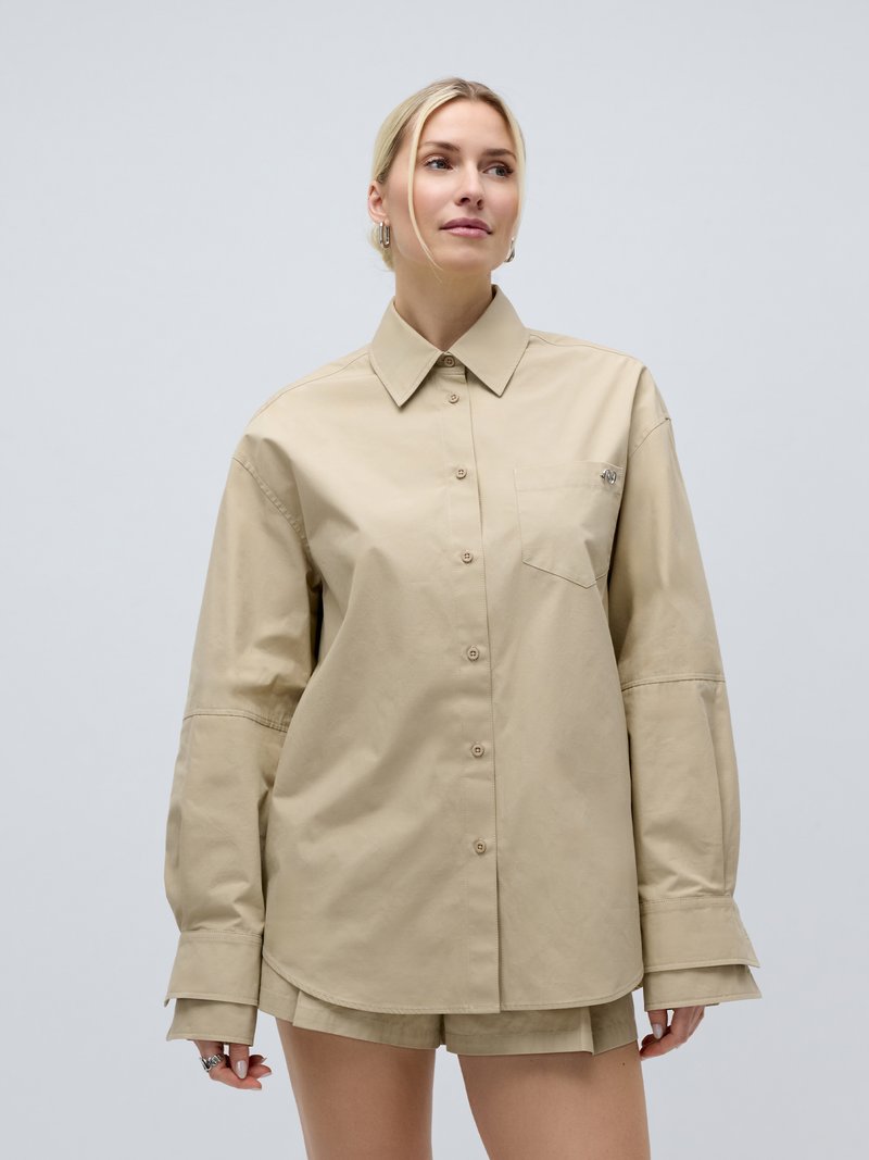 Γυναίκα με ξανθά μαλλιά που φοράει μια υπερμεγέθη μπεζ πουκάμισο με κουμπιά και ταιριαστό σορτς, στέκεται μπροστά σε απλό φωτεινό φόντο.