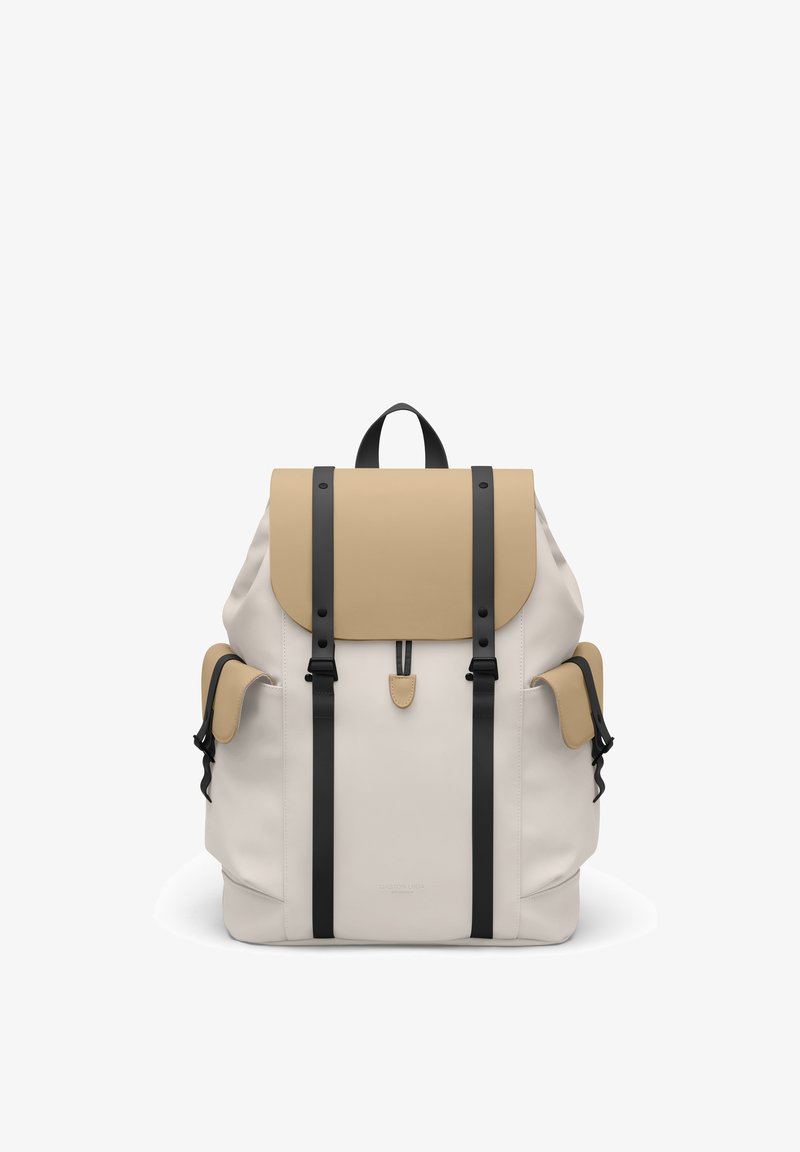 Sac à dos beige et crème avec bretelles noires, fermeture à rabat, deux poches latérales et poignée supérieure, présenté sur un fond blanc.
