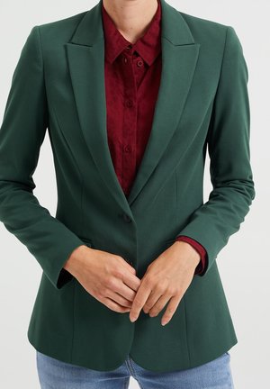 Personne boutonnant un bouton sur un blazer vert foncé par-dessus une chemise bordeaux, portant un jean bleu clair, sur un fond uni.